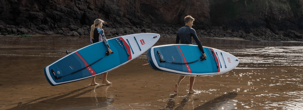 2 people carrying their paddleboards across the beach into the ocean