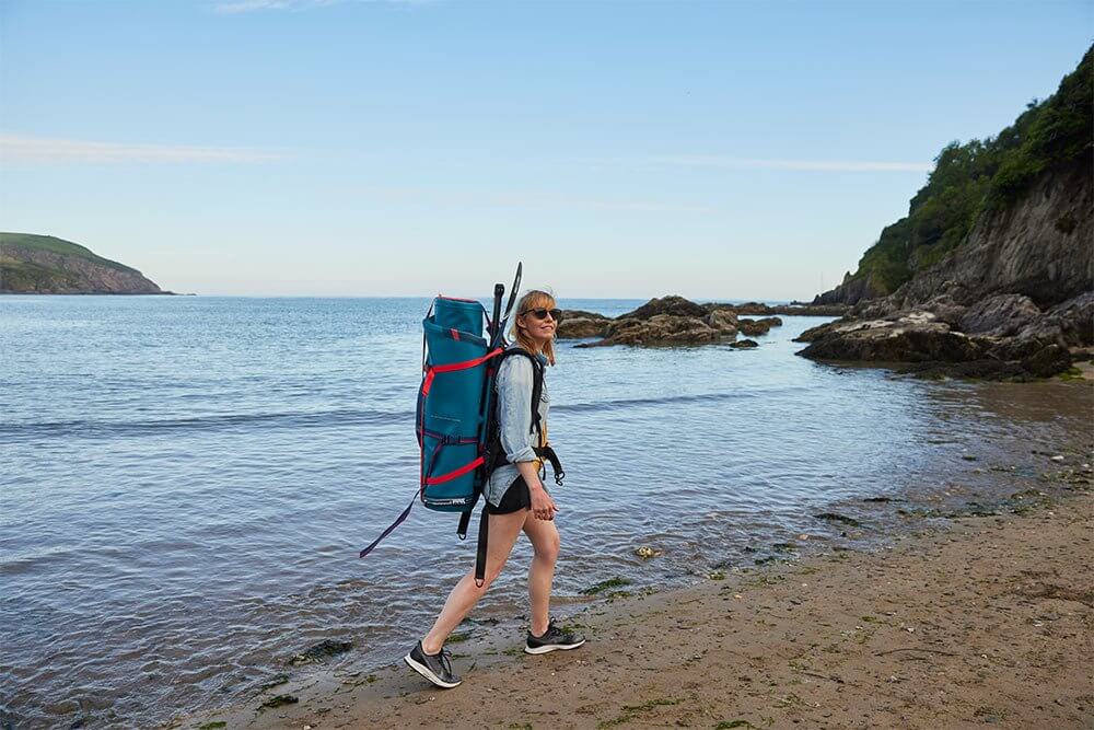 woman walking along the beach carrying a paddle board bag