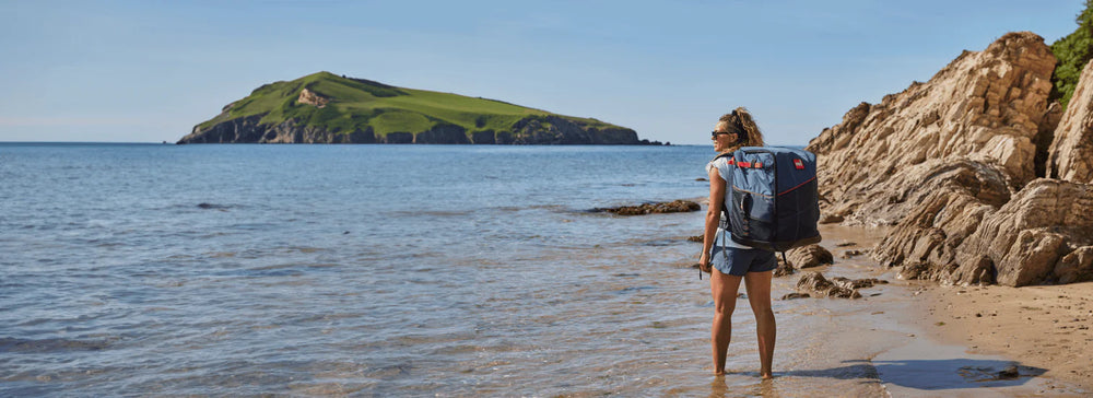 A woman walking on the beach, looking out to sea, wearing a red backpack