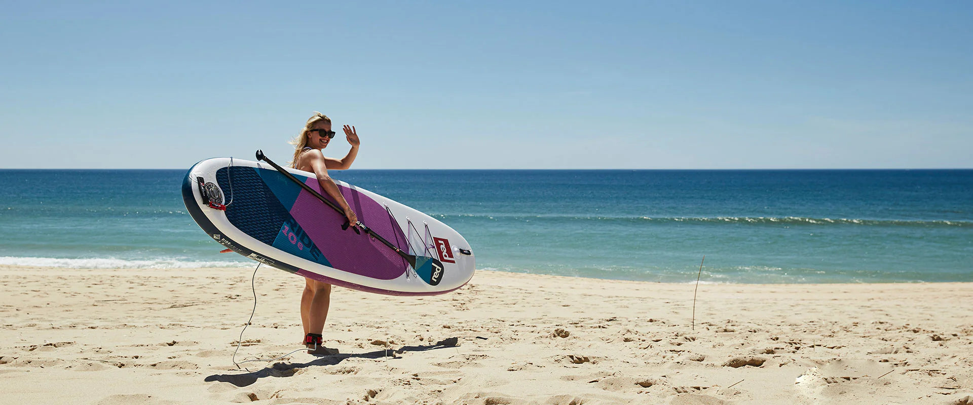 A woman on the beach, carrying her paddleboard, waving