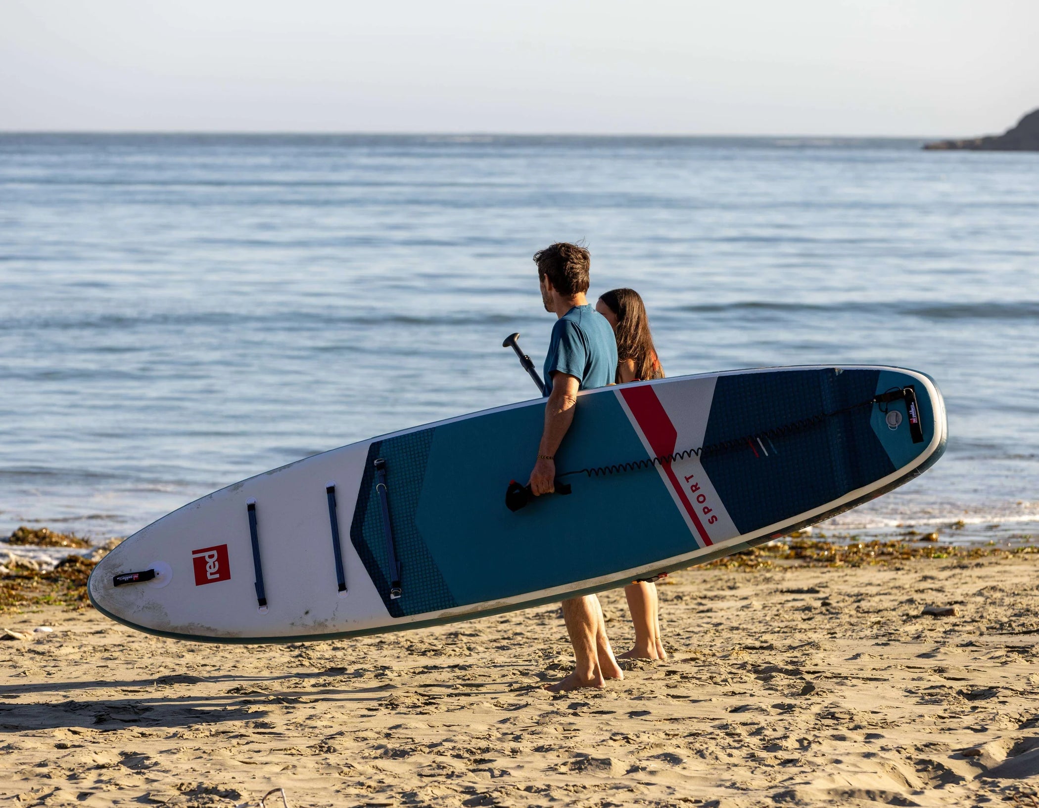 A man and woman walking along the beach with the man carrying a paddleboard