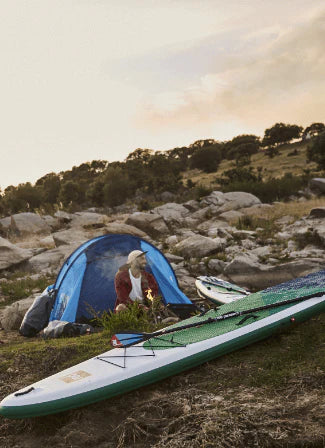 A man camping, sat in front of a fire with a paddleboard placed in the foreground