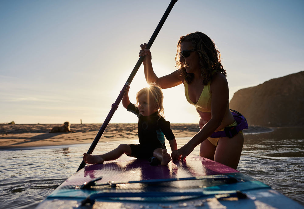 A mum holding her child on her paddleboard while she paddles in the sea on a sunny day