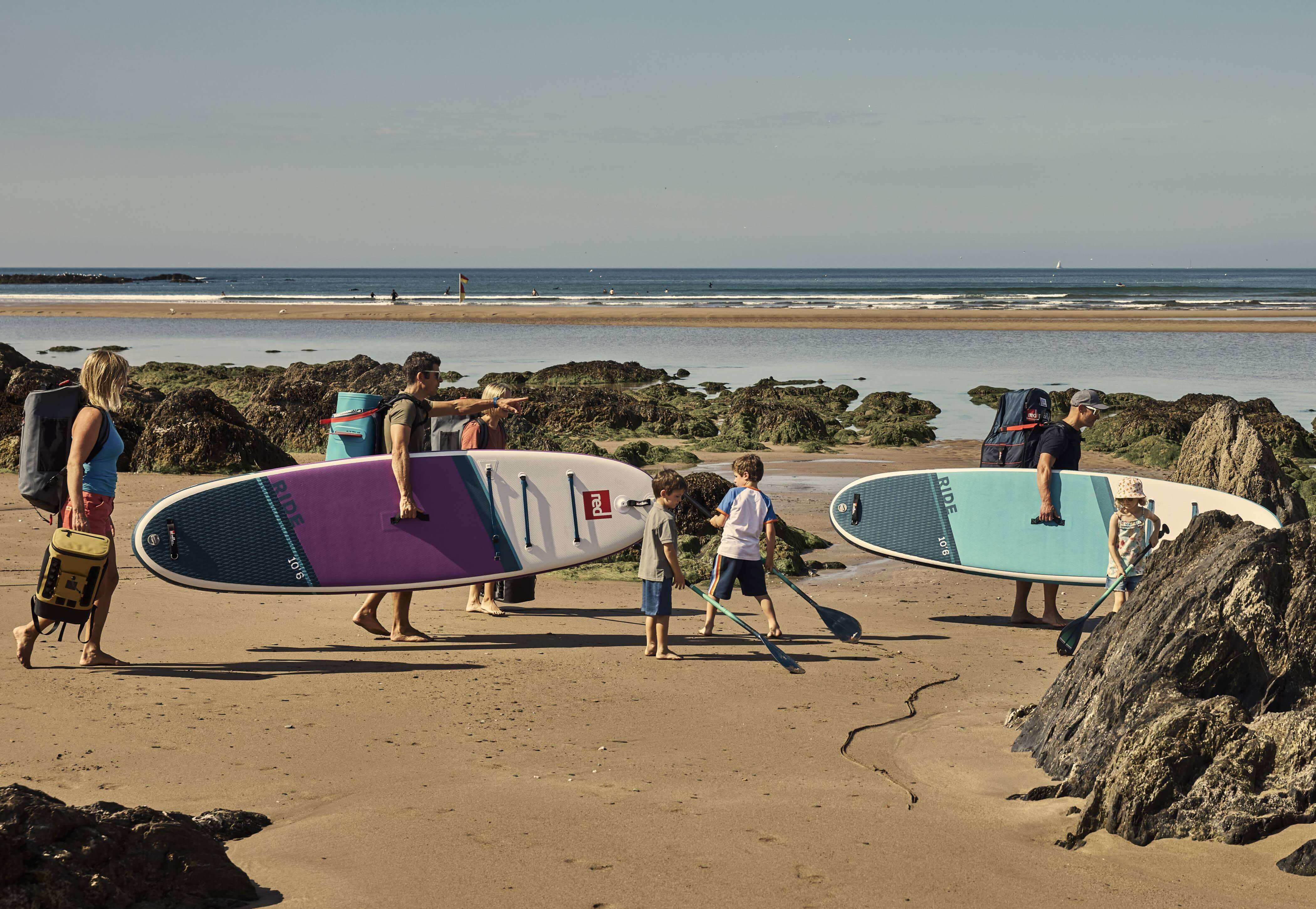 Family walking on beach, carrying Red Ride paddle boards, cooler backpacks, kit bag and paddles.