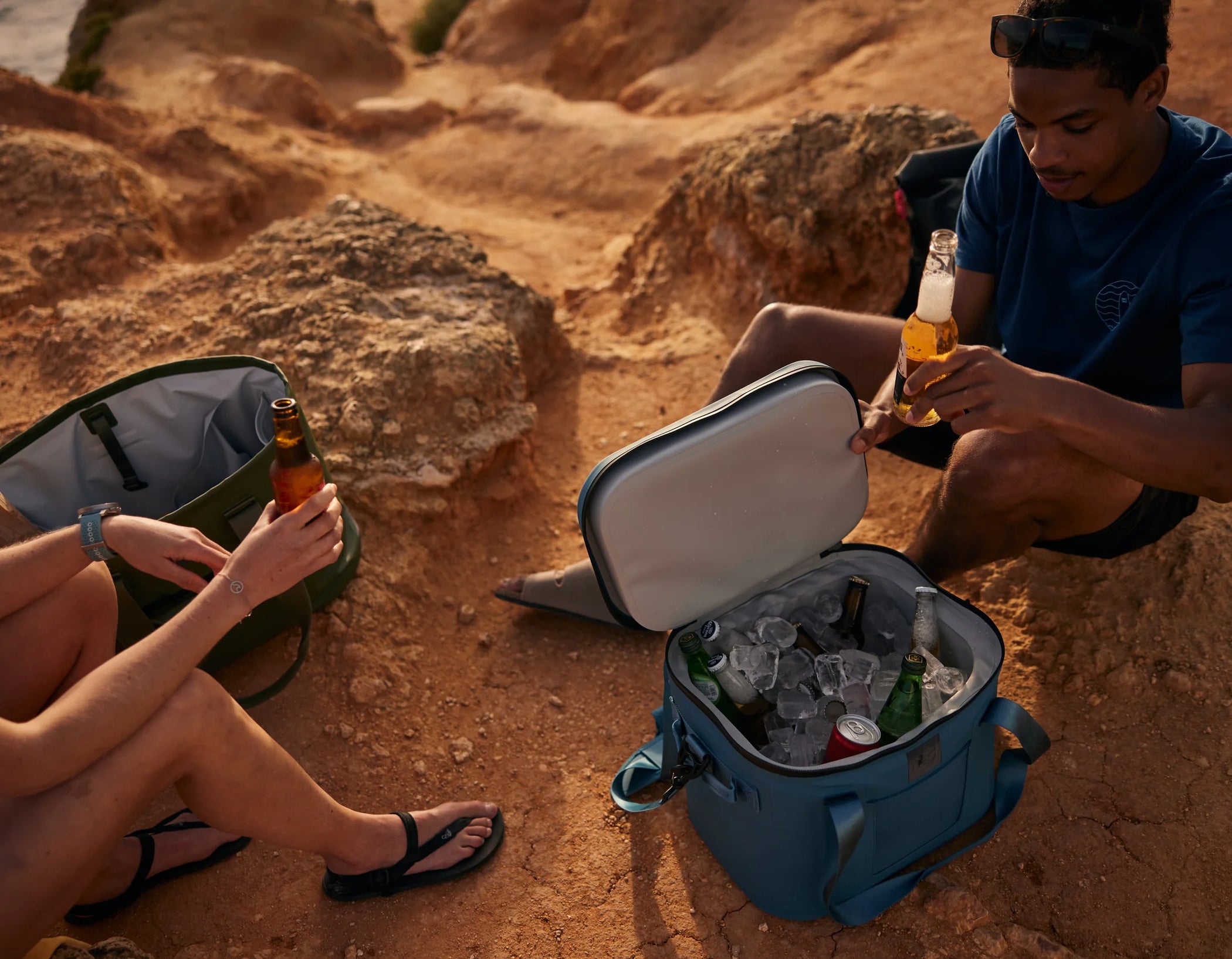 A cooler bag filled with drinks on the beach, 2 people sat around the bag