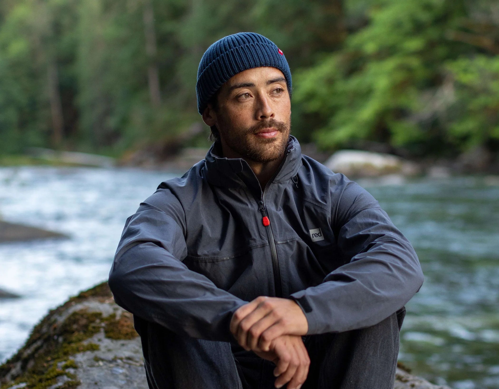 A man wearing a navy flow beanie, sat on a rock beside a river