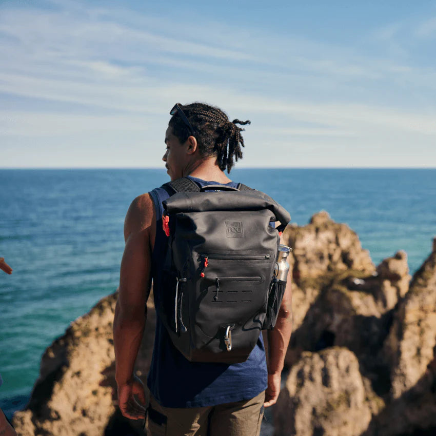 A man wearing the red backpack, looking out to sea